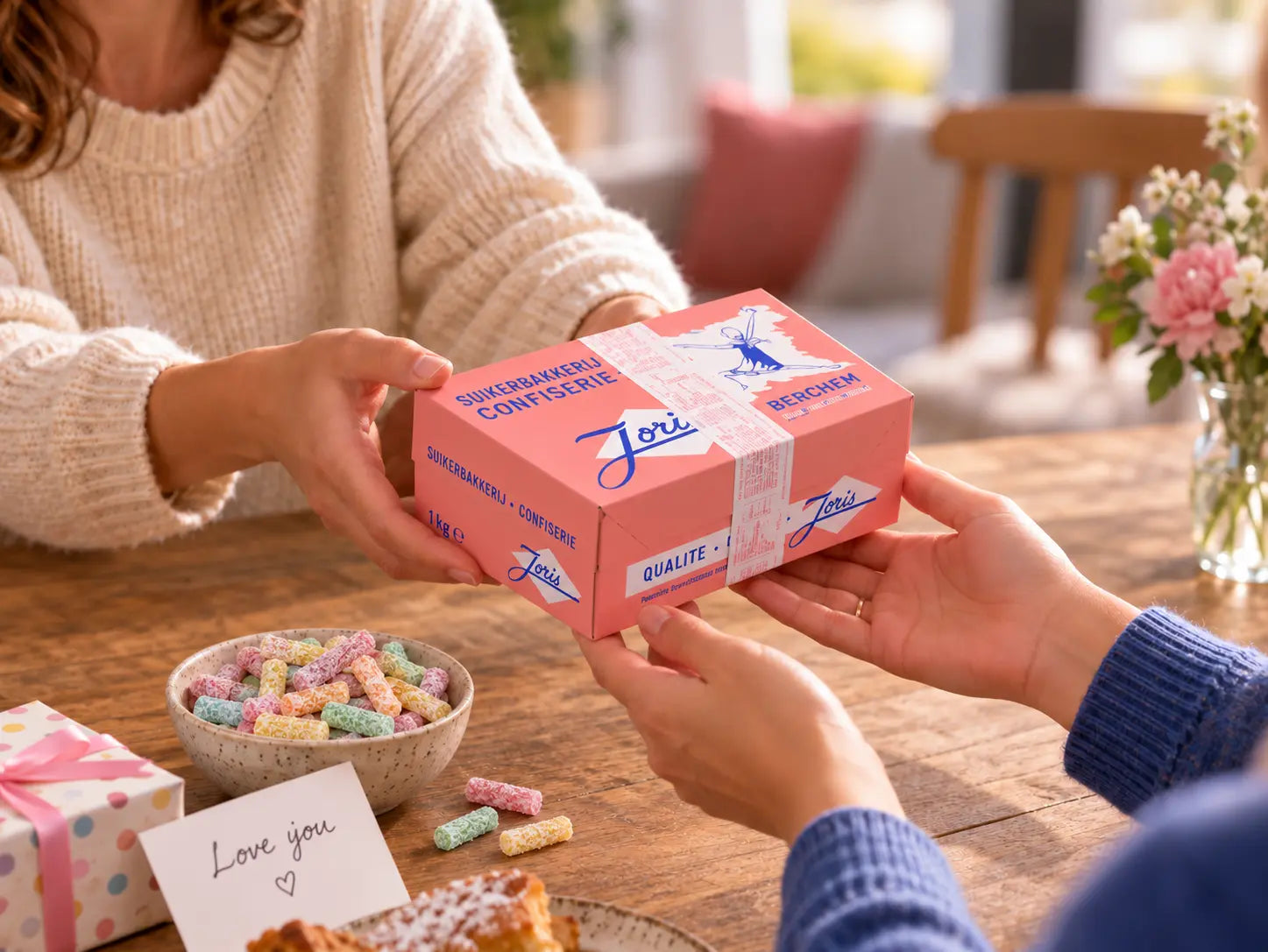 Twee personen geven elkaar een roze Joris Sweets snoepdoos van 1 kg aan tafel, met kleurrijke snoepjes, bloemen en een kaartje met "Love you".